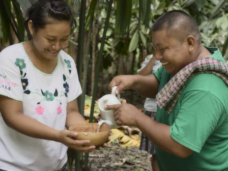 Two people outdoors, one pouring a white liquid into a bowl held by the other, both smiling and dressed casually.