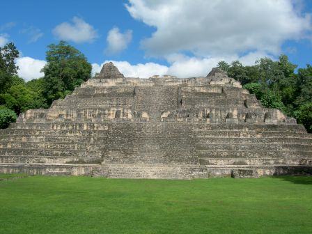 The image shows an ancient stone pyramid with a series of steps and terraces, surrounded by lush greenery under a partly cloudy sky.