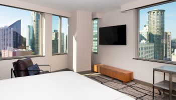 A modern bedroom with large windows offering a city view, wall-mounted TV, armchair, small table, and a low wooden cabinet.