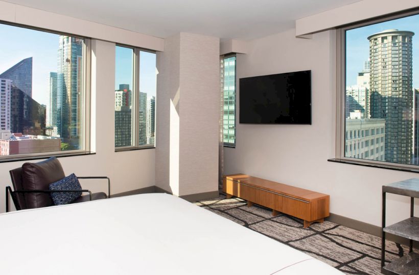 A modern bedroom with large windows offering a city view, wall-mounted TV, armchair, small table, and a low wooden cabinet.
