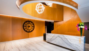 A modern lobby with a marble reception desk, wooden spiral staircase, wall clock, and hanging lights.