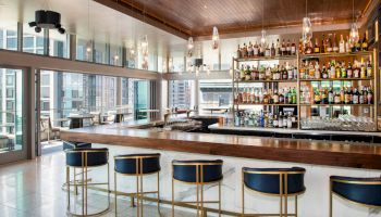 A modern bar with blue seats, a wooden countertop, and a large selection of bottles. Floor-to-ceiling windows show an urban view ending the sentence.