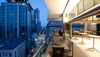 An upscale rooftop lounge in an urban setting with modern furniture and a view of tall buildings along a city street at twilight.