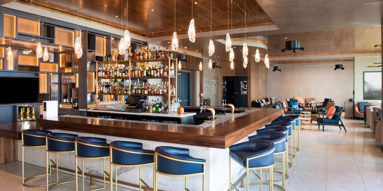 This image shows a modern bar with a long wooden counter, blue barstools, hanging lights, and a well-stocked liquor shelf, in a sleek setting.