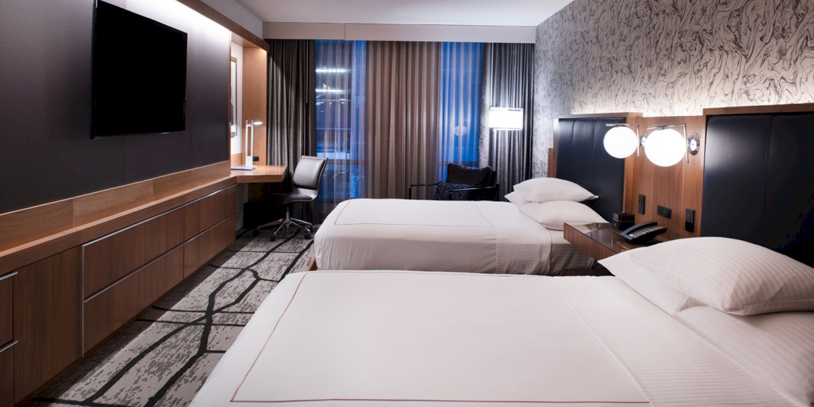 The image shows a modern hotel room with two neatly made beds, a desk, chair, TV, and stylish lighting.