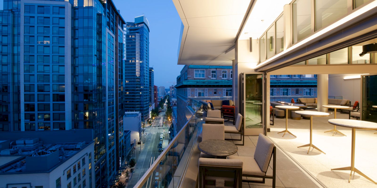 This image shows a modern urban balcony overlooking a cityscape with tall buildings at dusk. The balcony has chairs and tables for seating.