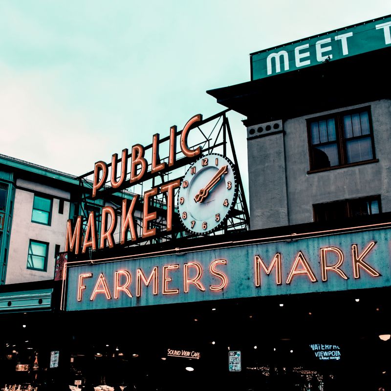 A historic public market sign with neon text reading "Public Market" and "Farmers Market," featuring a fish motif and a clock, on a cloudy day.