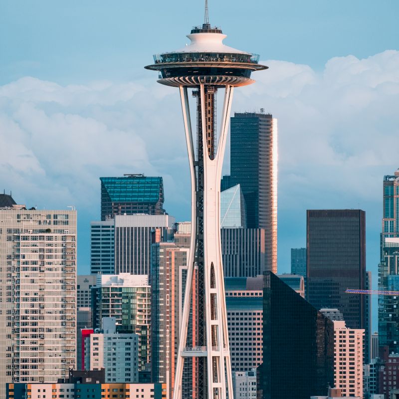 The image displays the skyline of a city with a prominent tower featuring a circular observation deck, set against a backdrop of various buildings and cloudy sky.