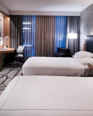 The image shows a modern hotel room with two neatly made beds, a wall-mounted TV, a work desk, and stylish decor and lighting.