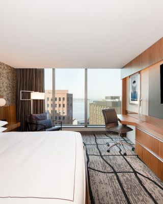 A modern hotel room with a large bed, desk, chair, flat-screen TV, and a window offering a city and water view.
