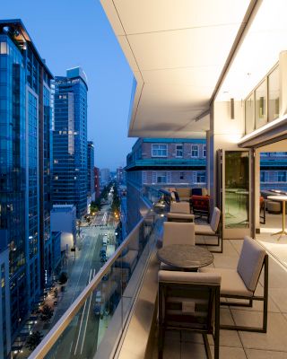 The image shows a modern urban balcony at twilight, overlooking a city street flanked by tall buildings. Tables and chairs are set up on the balcony.
