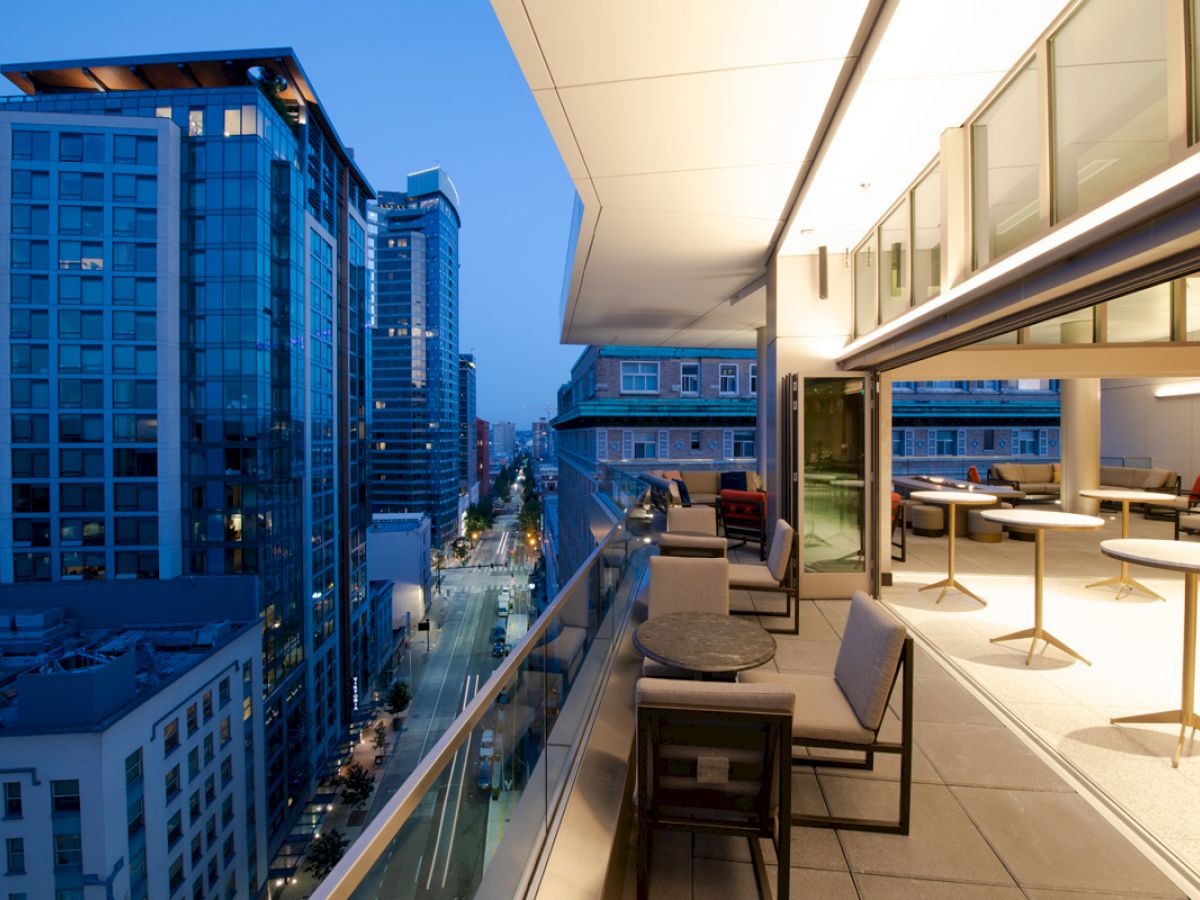 A modern balcony overlooking a city street with high-rise buildings, featuring seating and tables, at dusk or early evening.