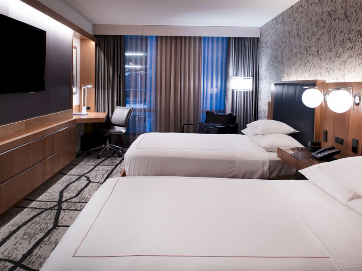 A modern hotel room with two beds, a TV, a desk, and a chair, featuring stylish decor and soft lighting.