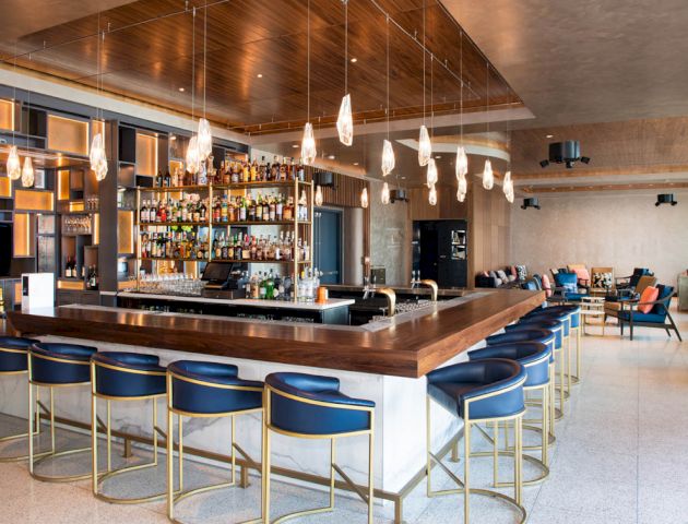 The image shows a modern bar with blue barstools, a well-stocked liquor shelf, hanging lights, and various lounge seating areas.