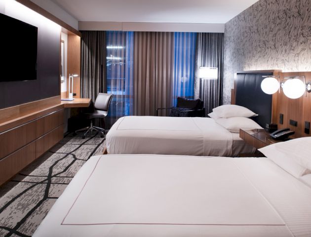 The image shows a modern hotel room with two neatly made beds, a flat-screen TV, a desk, and large windows with sheer curtains.