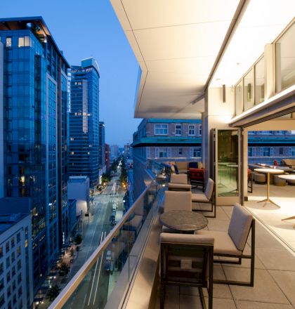 The image shows a modern balcony with tables and chairs overlooking a cityscape with tall buildings at dusk.