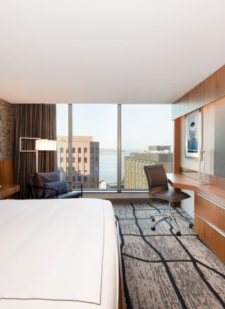 A modern hotel room with a large bed, wall-mounted TV, desk, and chair. The room has a city view through large windows.
