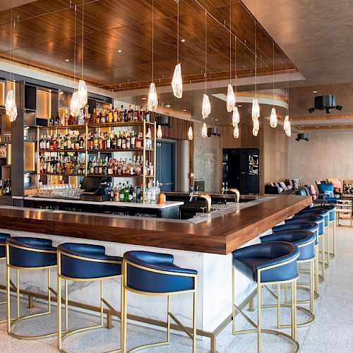 A modern bar features a large wooden countertop, elegant blue stools, hanging lights, and a well-stocked shelf of spirits.