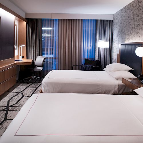 A modern hotel room with two neatly made beds, a wall-mounted TV, a desk with a chair, and contemporary decor and lighting.