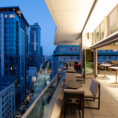 An urban setting with a modern high-rise building balcony featuring seating and tables; cityscape view with illuminated buildings at dusk.