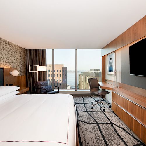 The image shows a modern hotel room with a large bed, wooden furnishings, a desk, a chair, and a view of buildings outside a large window.