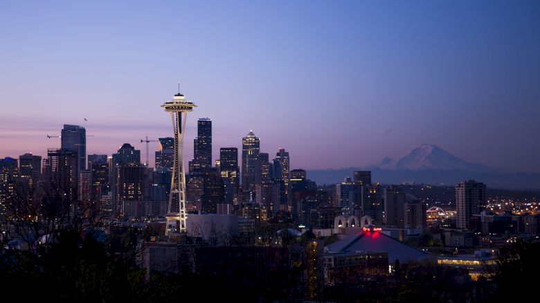 A panoramic view of a city skyline at dusk, featuring a distinctive tall tower and various buildings, with a mountain in the background.