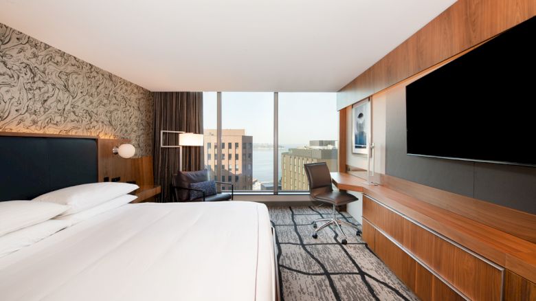 This image shows a modern hotel room with a king-sized bed, a desk, a large TV, and a view of city buildings from the window.