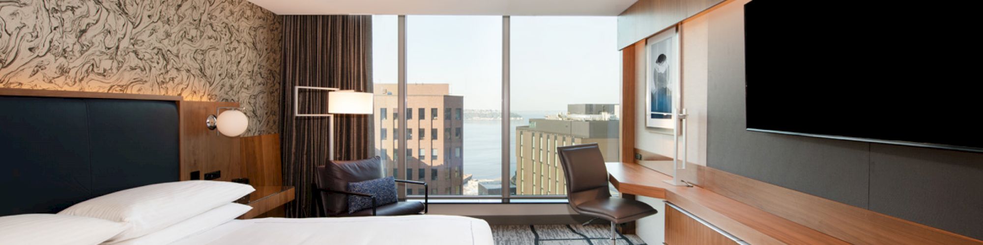 A modern hotel room with a large bed, desk, and chair, featuring a cityscape view through the window and a large flat-screen TV on the wall.