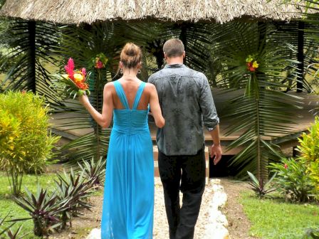 A couple is walking hand-in-hand towards a tropical gazebo, surrounded by lush greenery and vibrant flowers, in a serene outdoor setting.